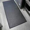 A Deckplate Anti-Fatigue Mat with a diamond plate pattern is placed on a speckled floor in a commercial kitchen, next to stainless steel appliances and worktops.