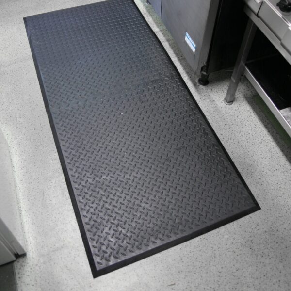 A Deckplate Anti-Fatigue Mat with a diamond plate pattern is placed on a speckled floor in a commercial kitchen, next to stainless steel appliances and worktops.
