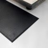 A Deckplate Anti-Fatigue Mat in black with a diamond plate pattern is placed on a smooth, light grey floor beside industrial equipment.