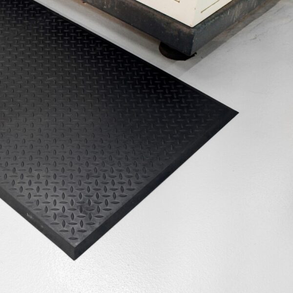 A Deckplate Anti-Fatigue Mat in black with a diamond plate pattern is placed on a smooth, light grey floor beside industrial equipment.