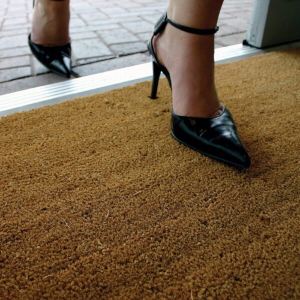 A close-up shows a person in black high heels stepping onto the Weather Resistant Entrance Mat at an entrance, with another foot behind on brick paving.