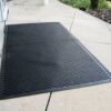 A Scraper Entrance Mat, in large black rubber with a textured pattern, is placed on a light-coloured tiled pavement by a building entrance and greenery.