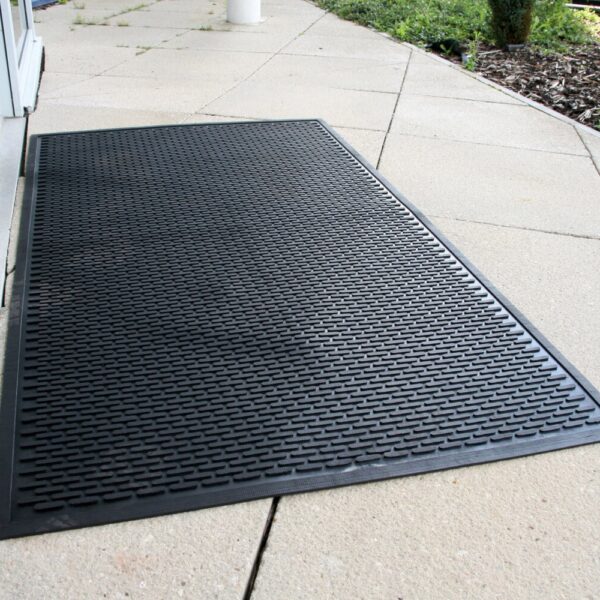 A Scraper Entrance Mat, in large black rubber with a textured pattern, is placed on a light-coloured tiled pavement by a building entrance and greenery.