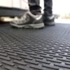 Close-up of the Scraper Entrance Mat with a textured, non-slip surface. In the background, a person in black trousers and grey trainers stands on the mat with their shoes slightly out of focus.