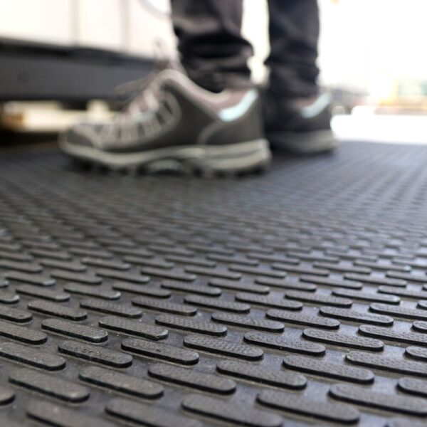 Close-up of the Scraper Entrance Mat with a textured, non-slip surface. In the background, a person in black trousers and grey trainers stands on the mat with their shoes slightly out of focus.