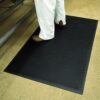 A person wearing white trousers and brown shoes stands on a Scraper Entrance Mat with a black textured surface, placed on a beige floor beside a metallic surface.