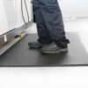 Someone stands on a Diamond Anti-Fatigue Mat—black and textured—wearing dark trousers and grey work shoes in a bright, clean indoor setting.