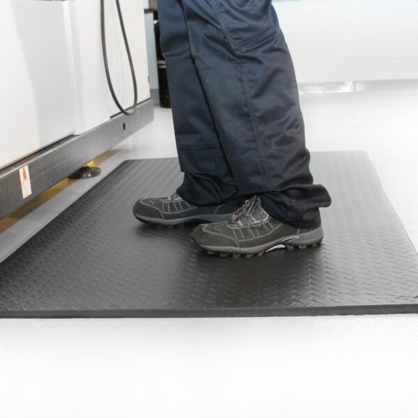 Someone stands on a Diamond Anti-Fatigue Mat—black and textured—wearing dark trousers and grey work shoes in a bright, clean indoor setting.