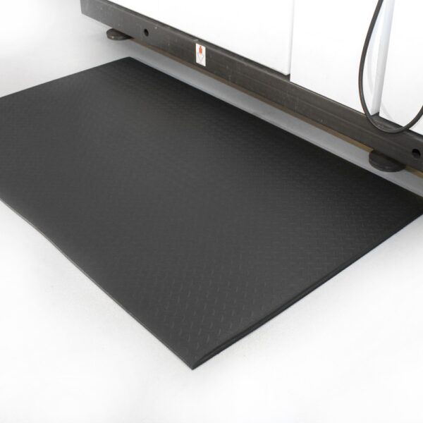 The Diamond Anti-Fatigue Mat, featuring a black diamond plate texture, is positioned on a white floor in front of a large industrial machine.