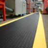 A Diamond Anti-Fatigue Mat with yellow safety edges runs along the floor of an industrial workspace, providing comfort and visibility near machinery and equipment.