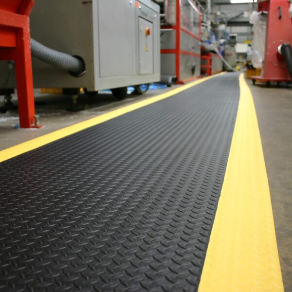 A Diamond Anti-Fatigue Mat with yellow safety edges runs along the floor of an industrial workspace, providing comfort and visibility near machinery and equipment.