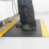A person in black trousers and shoes stands on a Premium Anti-Fatigue Mat with yellow edges, placed on a light-coloured floor beside a workstation.