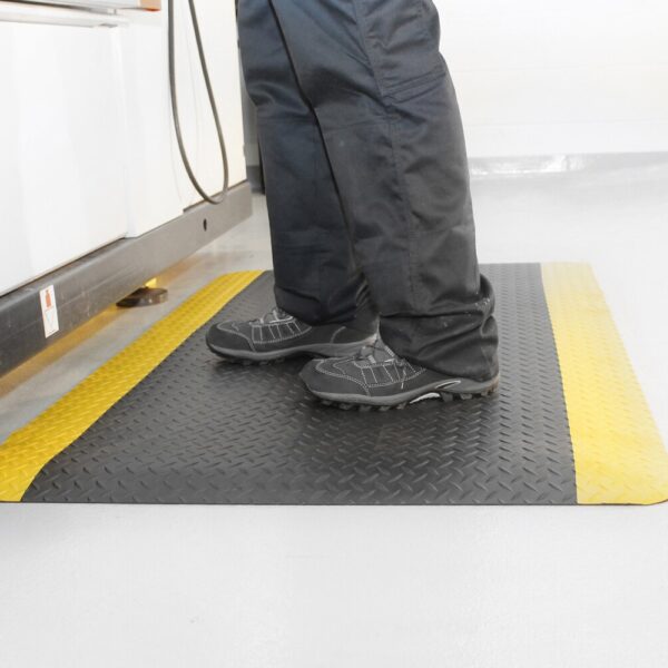 A person in black trousers and shoes stands on a Premium Anti-Fatigue Mat with yellow edges, placed on a light-coloured floor beside a workstation.