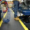 A person in jeans and tan work boots stands on the Premium Anti-Fatigue Mat, which features yellow lines, in a workshop near blue machinery and a coiled blue air hose hanging overhead.