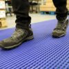 A person in black work shoes and trousers stands on a Self-Draining PVC Mat with a textured blue surface in an industrial setting.