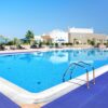 A self-draining PVC mat lies poolside by a large, clear blue outdoor swimming pool with a metal ladder, surrounded by loungers, parasols, palm trees, and a modern white building under the sunny sky.