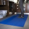 A person stands on a blue Self-Draining PVC Mat in a kitchen, surrounded by storage shelves, plastic containers, and stainless steel appliances.