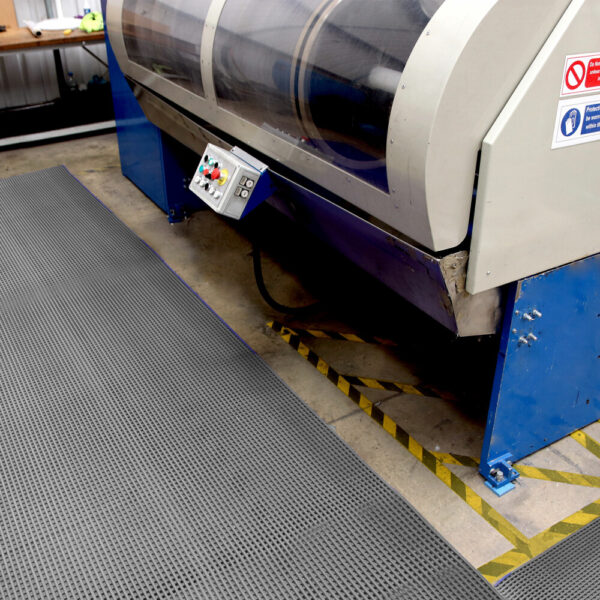 A Self-Draining PVC Mat lies near a large industrial machine on the factory floor, providing textured anti-fatigue support. The control panel and caution tape marking the machine's area are clearly visible.