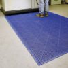 A person in work boots stands on an Interlocking Anti-Slip Leisure Tile, featuring a textured blue surface, placed on a light-coloured floor near industrial equipment.