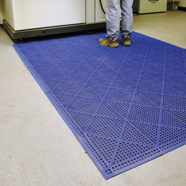 A person in work boots stands on an Interlocking Anti-Slip Leisure Tile, featuring a textured blue surface, placed on a light-coloured floor near industrial equipment.