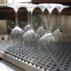 Six upside-down wine glasses are drying on an Interlocking Anti-Slip Leisure Tile in a kitchen or bar area, with stainless steel surfaces and a sink visible in the background.