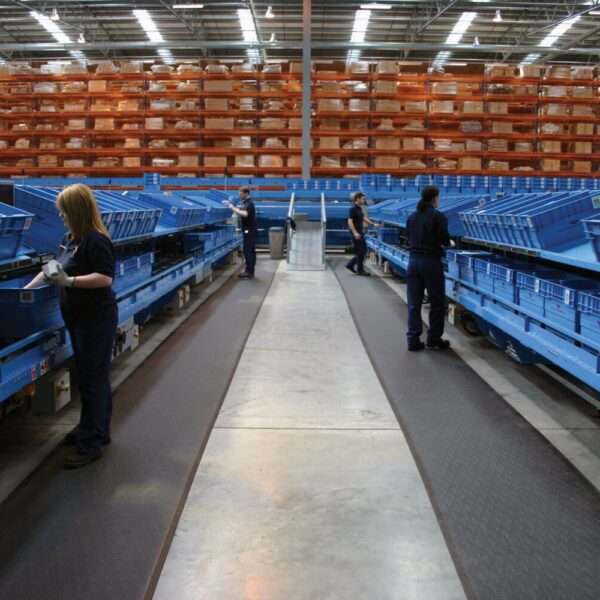 Workers stand beside blue conveyor belts on Textured Anti-Fatigue Mats, sorting parcels in a spacious, well-lit warehouse with high shelves, a wide central walkway, and neatly organised storage areas.
