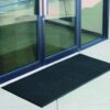 A Flexible Bristle Mat in black is placed outside a glass door with metal handles, reflecting its surroundings. The mat sits on a light-coloured, smooth floor surface.