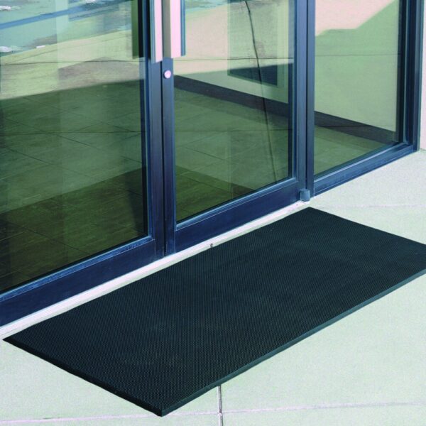 A Flexible Bristle Mat in black is placed outside a glass door with metal handles, reflecting its surroundings. The mat sits on a light-coloured, smooth floor surface.
