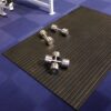 Two pairs of silver dumb-bells rest on a black Heavy-Duty Gym Mat, which is placed on a blue gym floor.