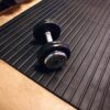 A single dumbbell rests on a heavy-duty gym mat with a black, ribbed surface, covering a wooden floor and creating a functional workout environment.