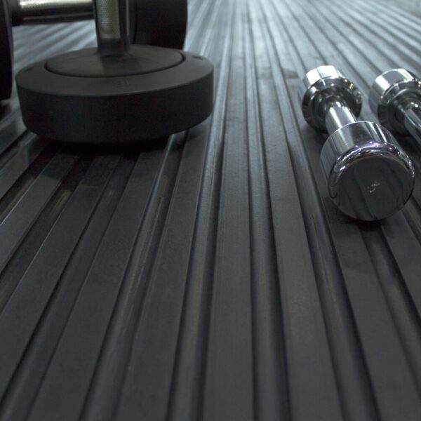 Three dumbbells sit on a Heavy-Duty Gym Mat’s ribbed black surface, with two chrome dumbbells on the right and a larger matte black one on the left, all viewed from a low angle.