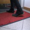 A person wearing black shoes and trousers steps onto the High-Performance Entrance Mat in red and black, placed on a tiled floor near a doorway.