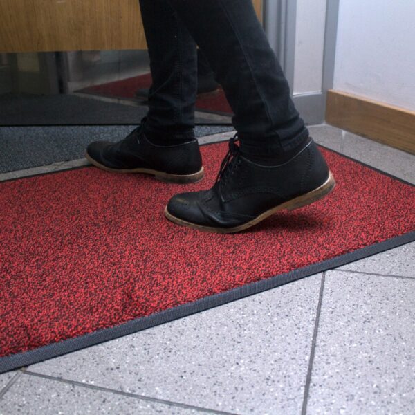 A person wearing black shoes and trousers steps onto the High-Performance Entrance Mat in red and black, placed on a tiled floor near a doorway.