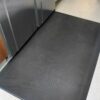 The Ultimate Anti-Fatigue Mat, in a large black size, is positioned in front of stainless steel appliances on a light speckled floor, perfect for kitchens or commercial areas.