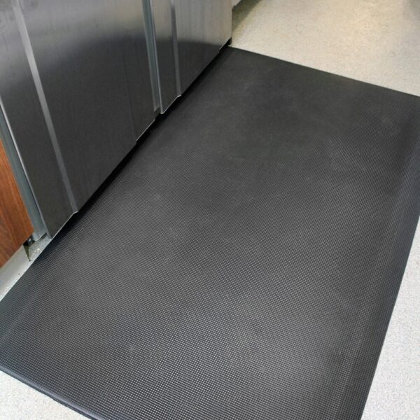 The Ultimate Anti-Fatigue Mat, in a large black size, is positioned in front of stainless steel appliances on a light speckled floor, perfect for kitchens or commercial areas.