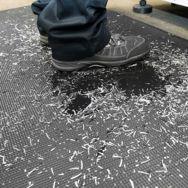 Close-up of a person wearing black work shoes standing on the Ultimate Anti-Fatigue Mat, which is covered with metal or plastic shavings in an industrial setting.