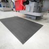 The Ultimate Anti-Fatigue Mat, black in colour, sits on a light workshop floor near industrial machines and a red tool chest, with small metal shavings scattered around it.