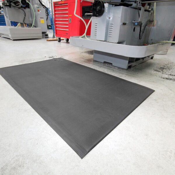 The Ultimate Anti-Fatigue Mat, black in colour, sits on a light workshop floor near industrial machines and a red tool chest, with small metal shavings scattered around it.