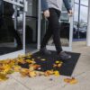 A person steps through a glass door onto an Octagonal Ring Mat, surrounded by yellow and orange autumn leaves near the entrance.