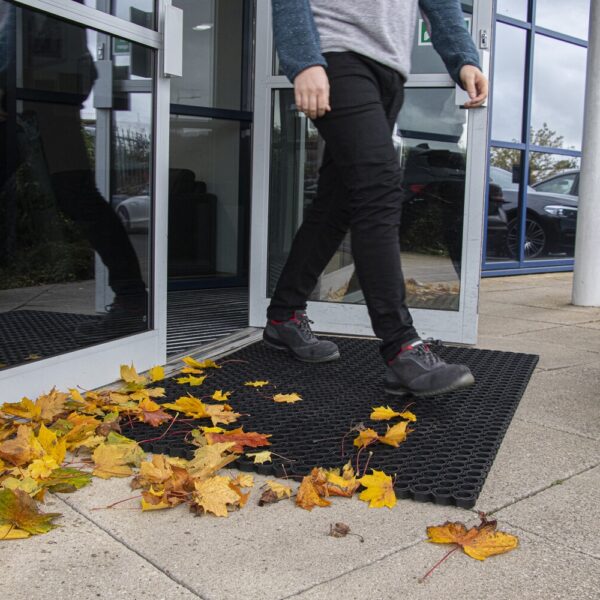 A person steps through a glass door onto an Octagonal Ring Mat, surrounded by yellow and orange autumn leaves near the entrance.