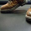 Close-up of a person wearing brown work boots standing on a Fine Ribbed Matting Roll, which features a black, ribbed anti-fatigue surface.