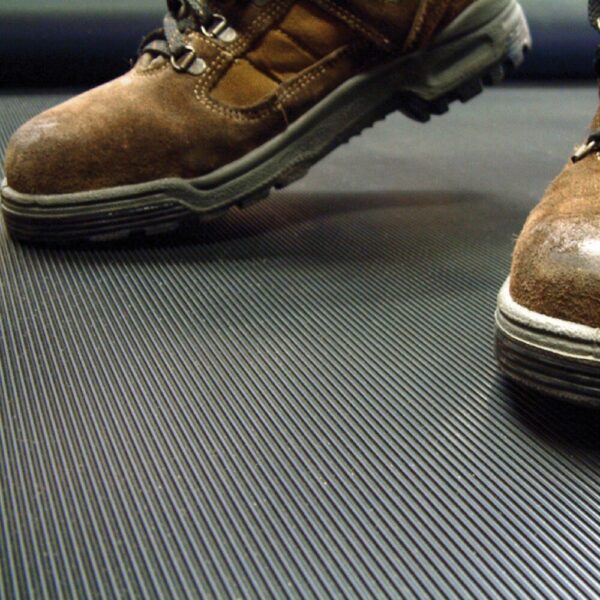 Close-up of a person wearing brown work boots standing on a Fine Ribbed Matting Roll, which features a black, ribbed anti-fatigue surface.