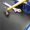 A close-up of a hammer, an adjustable spanner, and a spanner on Fine Ribbed Matting Roll, placed next to a blue metal vice on a dark workbench.