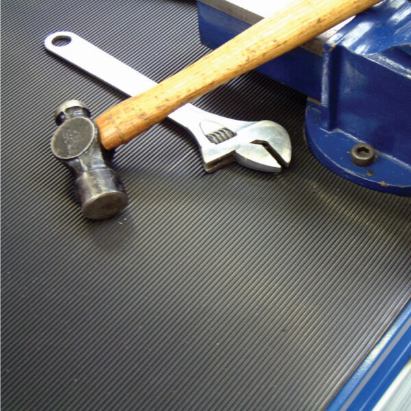 A close-up of a hammer, an adjustable spanner, and a spanner on Fine Ribbed Matting Roll, placed next to a blue metal vice on a dark workbench.