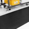 A Wide Ribbed Rubber Matting in black lies on a white floor beside a wheeled metal trolley with adjustable feet, supporting a large yellow bin.