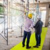 Two construction workers in hard hats stand on a yellow Construction Site Walkway Roll; one holds plans and points as they discuss progress near scaffolding and concrete structures.