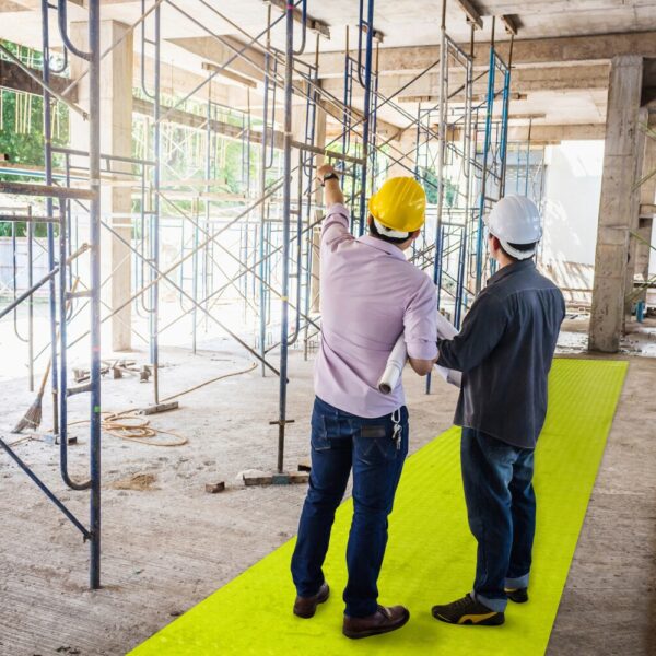 Two construction workers in hard hats stand on a yellow Construction Site Walkway Roll; one holds plans and points as they discuss progress near scaffolding and concrete structures.