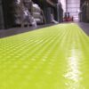 A close-up of the Construction Site Walkway Roll, a textured bright yellow safety mat installed on an industrial warehouse floor, with shelves and a forklift seen in the background.