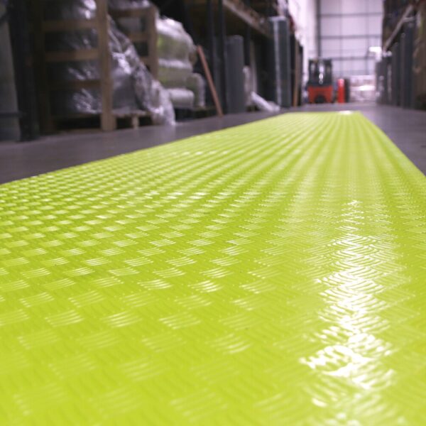A close-up of the Construction Site Walkway Roll, a textured bright yellow safety mat installed on an industrial warehouse floor, with shelves and a forklift seen in the background.