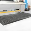 A Nitrile Top Interlocking Mat with holes in black is placed on a white floor in front of industrial machinery, with tools and boxes visible in the background.